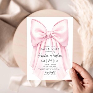 Op de afbeelding: Een uitnodiging voor een babyshower met een grote, aquarel roze strik. De uitnodiging bevat de tekst "Join us for a Baby Shower in Honor of Sophia Hudson" en evenementdetails. De uitnodiging wordt door een hand vastgehouden.