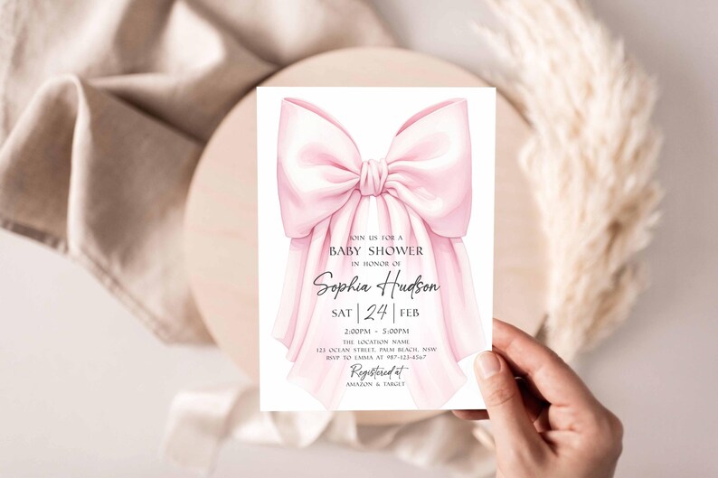 Op de afbeelding: Een uitnodiging voor een babyshower met een grote, aquarel roze strik. De uitnodiging bevat de tekst "Join us for a Baby Shower in Honor of Sophia Hudson" en evenementdetails. De uitnodiging wordt door een hand vastgehouden.