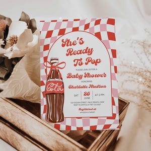 May include: A baby shower invitation with a retro design. The invitation features a Coca-Cola bottle illustration and the text "She's Ready To Pop". The background has a red and pink checkered pattern. The invitation is in a wooden crate with flowers.