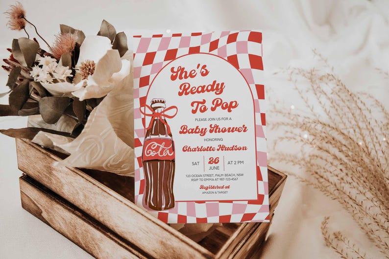 May include: A baby shower invitation with a retro design. The invitation features a Coca-Cola bottle illustration and the text "She's Ready To Pop". The background has a red and pink checkered pattern. The invitation is in a wooden crate with flowers.
