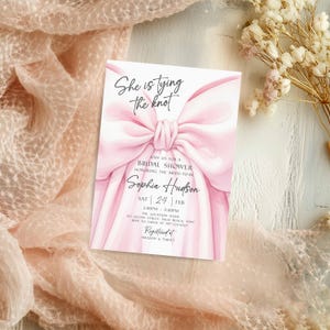 K&ouml;nnte beinhalten: Eine rosa Brautparty-Einladung mit einem gro&szlig;en Schleifendesign. Der Text "She is tying the knot" steht oben, mit Details f&uuml;r Sophias Dusche darunter. Die Einladung liegt auf einer strukturierten Oberfl&auml;che.