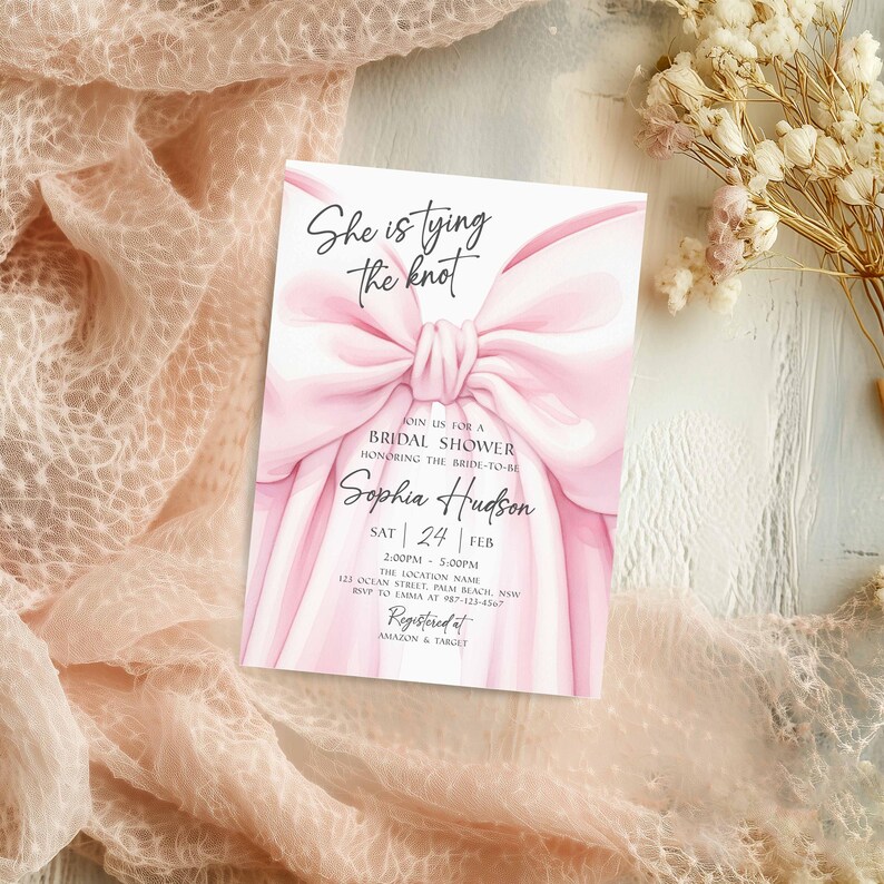 K&ouml;nnte beinhalten: Eine rosa Brautparty-Einladung mit einem gro&szlig;en Schleifendesign. Der Text "She is tying the knot" steht oben, mit Details f&uuml;r Sophias Dusche darunter. Die Einladung liegt auf einer strukturierten Oberfl&auml;che.