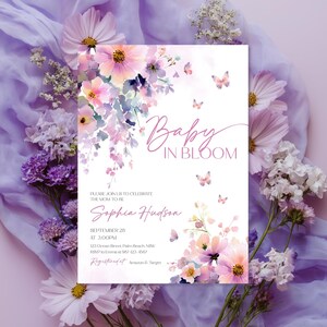 Może przedstawiać: Zaproszenie na baby shower ze słowami "Baby in Bloom" w eleganckim skrypcie. Zaproszenie zawiera akwarelowe kompozycje kwiatowe w odcieniach r&oacute;żu, fioletu i ż&oacute;łci, z delikatnymi akcentami motyli. Zaproszenie zawiera imię Sophia Hudson i szczeg&oacute;ły wydarzenia.