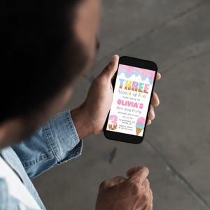 May include: A person holding a smartphone displaying a birthday party invitation. The invitation features the text "Three Scoops of Fun" and "Olivia's Birthday Party" in pink and blue colors, with ice cream cone illustrations.