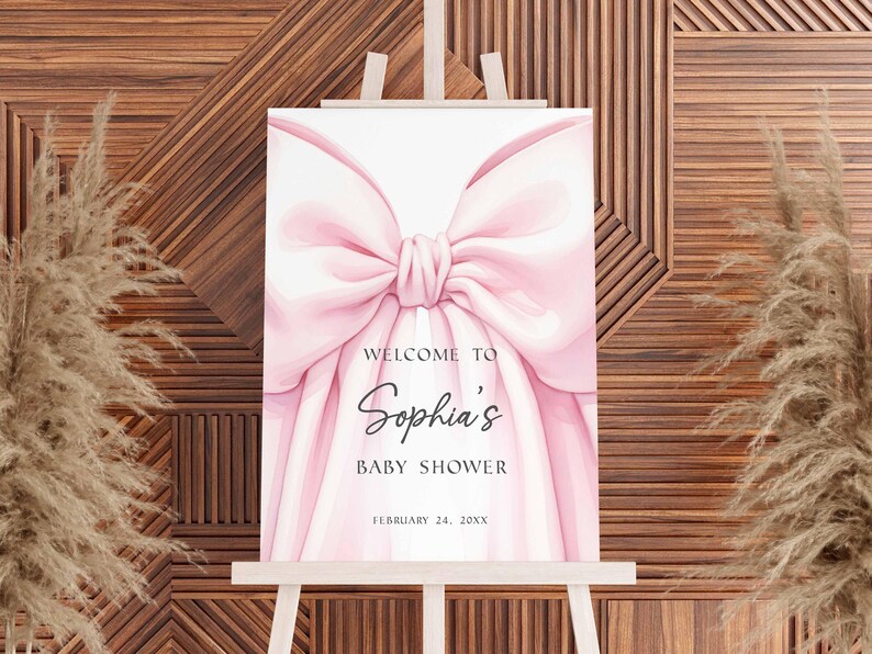 May include: A baby shower sign on a wooden easel. The sign features a large pink bow design with the words "Welcome to Sophia's Baby Shower" and the date February 24, 20XX.