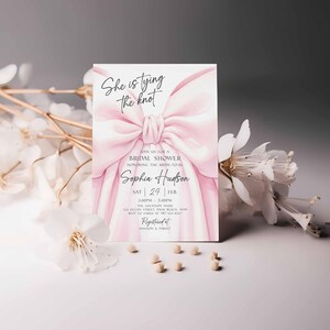 K&ouml;nnte beinhalten: Eine Einladung zur Brautparty mit einem rosa Aquarell-Schleifendesign. Die Einladung enth&auml;lt den Text "She is tying the knot" und Details zur Veranstaltung. Wei&szlig;e Blumen und kleine Perlen befinden sich im Vordergrund.