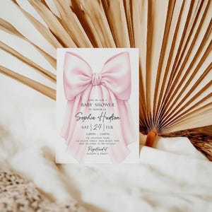 Op de afbeelding: Een uitnodiging voor een babyshower met een grote roze strik. De uitnodigingstekst bevat de woorden "Ann us for a Baby Shower in Honor of Sophia Hudson", samen met de datum en tijd. De achtergrond toont gedroogde palmbladeren.