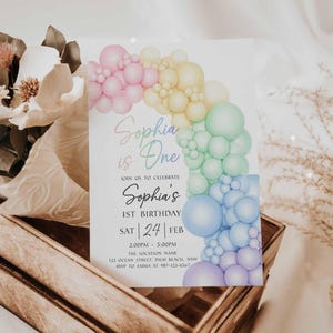May include: A birthday invitation with a rainbow balloon arch design. The invitation reads "Sophia is One" and includes details for the 1st birthday celebration, including the date, time, and location. The invitation is set in a wooden crate with flowers.