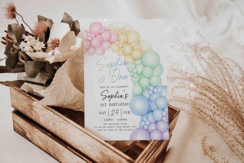 May include: A birthday invitation with a rainbow balloon arch design. The invitation reads "Sophia is One" and includes details for the 1st birthday celebration, including the date, time, and location. The invitation is set in a wooden crate with flowers.