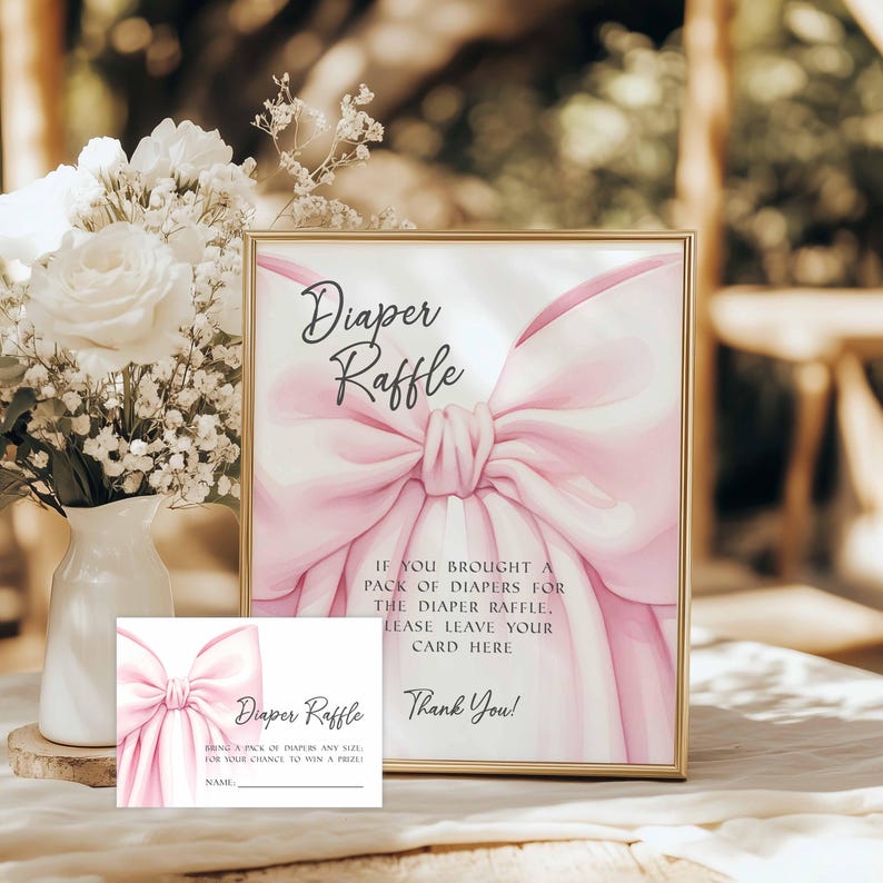 May include: A gold-framed sign and a card with the text "Diaper Raffle" and a pink bow illustration. The sign reads "If you brought a pack of diapers for the diaper raffle, please leave your card here. Thank You!" A white vase with white flowers is in the background.