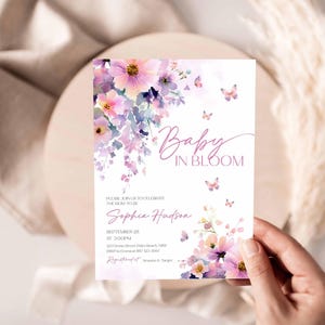 Może przedstawiać: Karta zaproszenia na baby shower z kwiatowym wzorem akwarelowym w odcieniach r&oacute;żu i fioletu. Karta zawiera tekst "Baby in Bloom" i szczeg&oacute;ły wydarzenia. Motyle i kwiaty zdobią kartę, trzymaną w dłoni.