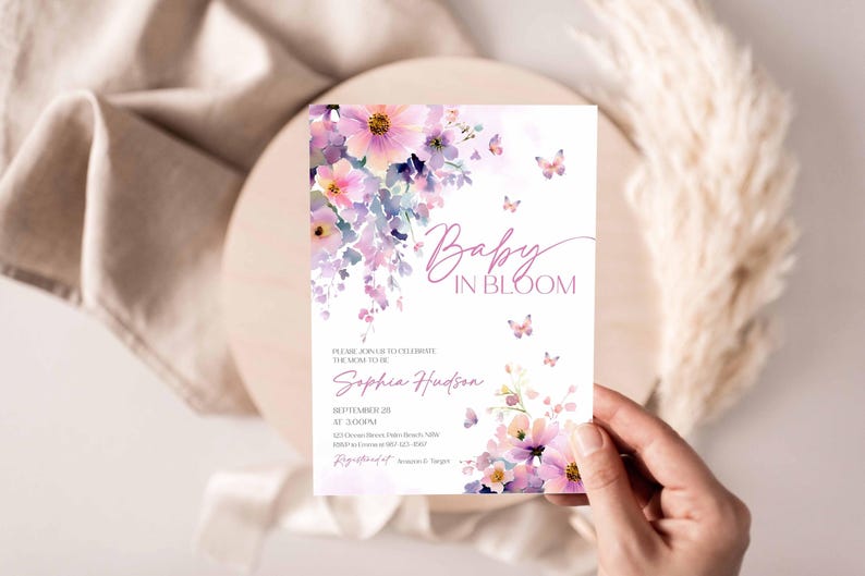 Może przedstawiać: Karta zaproszenia na baby shower z kwiatowym wzorem akwarelowym w odcieniach r&oacute;żu i fioletu. Karta zawiera tekst "Baby in Bloom" i szczeg&oacute;ły wydarzenia. Motyle i kwiaty zdobią kartę, trzymaną w dłoni.