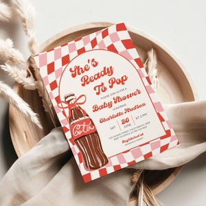 May include: A baby shower invitation with a retro design. The invitation features a red and pink checkered border and a vintage cola bottle illustration. Text includes "She's Ready To Pop" and details for the event.