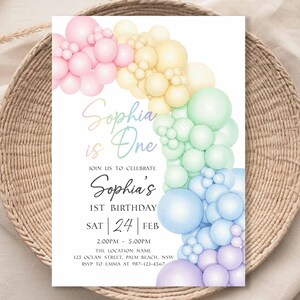 May include: A birthday invitation with a rainbow balloon arch design. The text "Sophia is One" is displayed in a script font. Additional text includes the date, time, and location for the celebration.