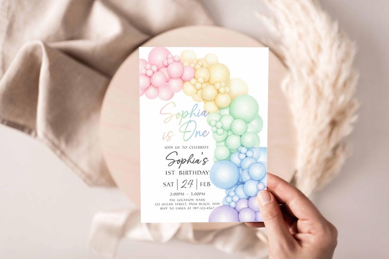 May include: A birthday invitation featuring a colorful balloon arch in pastel shades of pink, yellow, green, and blue. The text "Sophia is One" is displayed in elegant script, along with event details. The invitation is held by a hand.