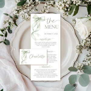 May include: Elegant menu and place card set with a soft green ribbon design. The menu features the text "the MENU" and a list of courses and dishes. The place card has the name "Charlotte". The design is set on a white plate with floral accents.
