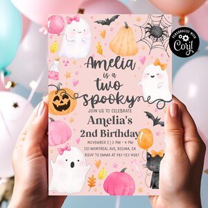 May include: A pink Halloween-themed invitation with the text "Amelia is a two spookyne". The invitation features illustrations of ghosts, pumpkins, bats, and a black cat. The invitation is held by a person.