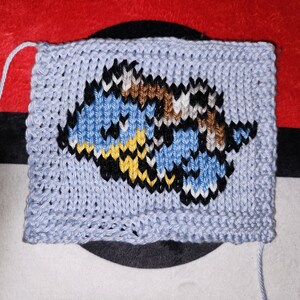 May include: A knitted square with a blue, yellow, black, white, and brown pixelated image of a cartoon character. The square is made of light blue yarn.