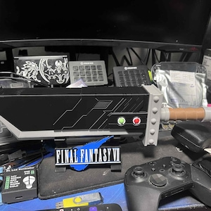 May include: A black and gray replica Buster Sword from Final Fantasy VII, with a brown handle and green and red buttons. The sword rests on a stand with the game's logo. A black game controller is in the foreground.