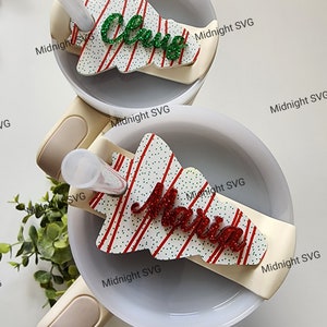 May include: Two white mugs with a red and white striped Christmas tree design. The tree has green glitter lettering that says "Claus" and "Merry".