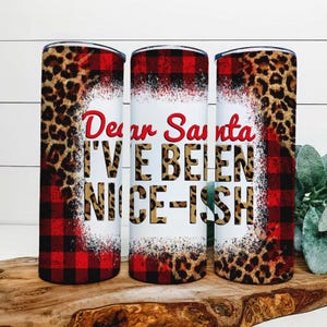 May include: Three stainless steel tumblers with a festive design. The tumblers feature a mix of red buffalo plaid, leopard print, and the text "Dear Santa I've Been Nice-ish" in red and leopard print.