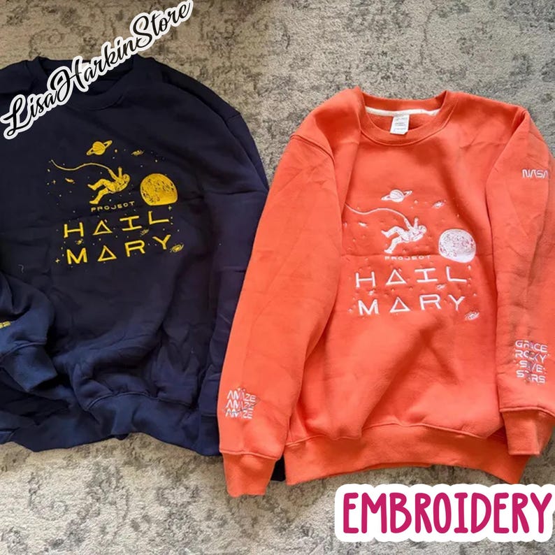 May include: A dark navy blue sweatshirt and an orange sweatshirt, both featuring a space-themed graphic with an astronaut, planets, and the text Project Hail Mary. The orange sweatshirt includes NASA branding and additional text on the sleeve.