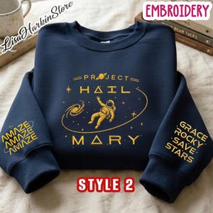 May include: A navy blue crewneck sweatshirt featuring gold embroidery. The chest displays an astronaut floating in space with the text PROJECT HAIL MARY. The left sleeve reads AMAZE repeated, and the right sleeve reads GRACE ROCKY SAVE STARS.