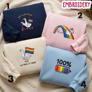 May include: Four sweatshirts with embroidered designs. One is navy blue with a goose on a scooter and the text "Off to Be Proud." Another is pink with a rainbow and a goose, with the text "Just a Proud Goose." A cream sweatshirt has a goose with a rainbow flag and the text "We Pride at Dawn." The last is light blue with a rainbow battery and the text "100%."