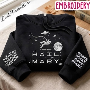 May include: A black hooded sweatshirt featuring a white graphic of an astronaut floating in space with the text Project Hail Mary. The left sleeve displays the word Amaze repeatedly, while the right sleeve features the text Grace Rocky Save Stars.