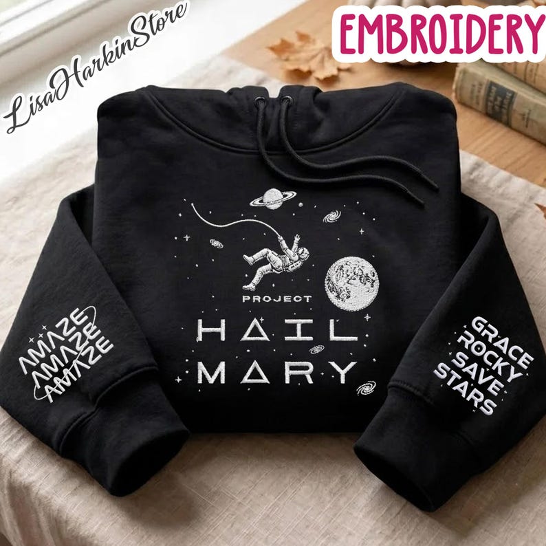 May include: A black hooded sweatshirt featuring a white graphic of an astronaut floating in space with the text Project Hail Mary. The left sleeve displays the word Amaze repeatedly, while the right sleeve features the text Grace Rocky Save Stars.