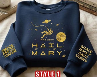 May include: A navy blue crewneck sweatshirt featuring gold embroidery. The front displays an astronaut floating in space, a planet, and the text "PROJECT HAIL MARY". The left sleeve reads "AMAZE" repeatedly, and the right sleeve reads "GRACE ROCKY SAVE STARS".