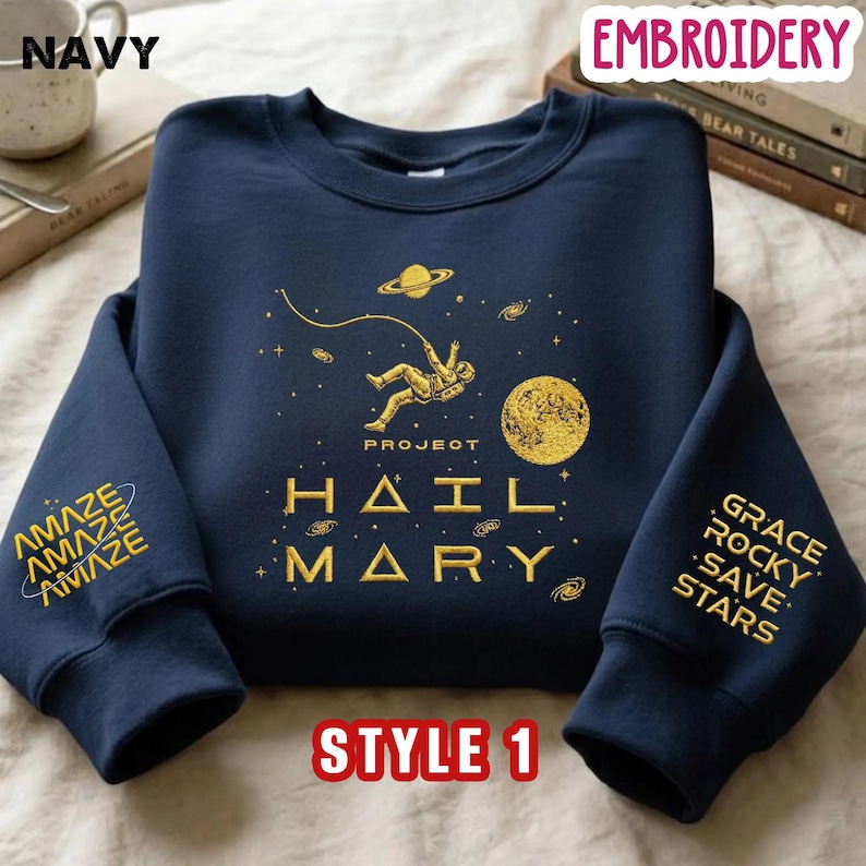 May include: A navy blue crewneck sweatshirt featuring gold embroidery of an astronaut floating in space with a planet, stars, and the text PROJECT HAIL MARY. The left sleeve displays the word AMAZE, and the right sleeve lists GRACE ROCKY SAVE STARS.