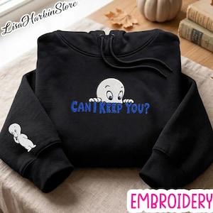 Peut inclure: Un sweat à capuche noir avec une broderie représentant un fantôme blanc qui regarde par-dessus le texte "CAN I KEEP YOU?" en bleu. Un petit fantôme blanc est brodé sur la manche droite. Le sweat possède des cordons de serrage noirs assortis.