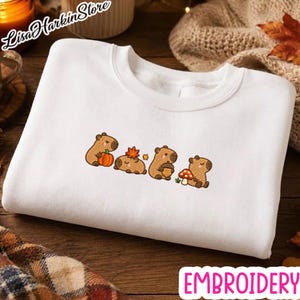 May include: A white crewneck sweatshirt featuring an embroidered design of four brown capybaras. The capybaras are depicted interacting with autumn-themed items including an orange pumpkin, a red maple leaf, a brown acorn, and a red mushroom.