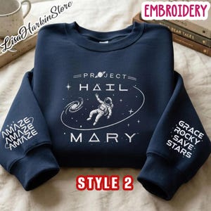 May include: A navy blue crewneck sweatshirt featuring white embroidery. The chest displays an astronaut floating in space with the text "PROJECT HAIL MARY" and a planet. The left sleeve reads "AMAZE" and the right sleeve reads "GRACE ROCKY SAVE STARS".