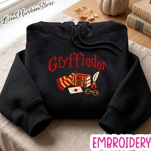 May include: A black hooded sweatshirt featuring red embroidered text that reads Gryffindor. Below the text is an embroidered graphic of a red and yellow striped scarf, a stack of books, a quill in an inkwell, a wand, and round glasses.