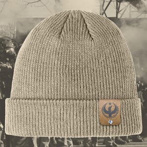 May include: A tan ribbed knit beanie with a folded brim. A small, rectangular brown leather patch is sewn on the front, featuring a dark blue emblem of a bird with outstretched wings. The patch is secured with a silver rivet.
