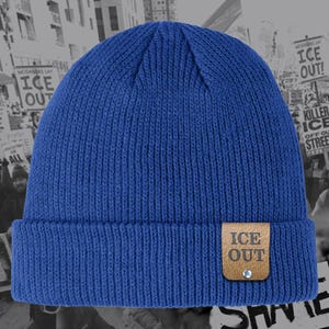 May include: A blue knit beanie with a folded brim. A small, brown leather tag is attached to the brim, with the words "ICE OUT" printed in orange. The beanie is a solid color and has a ribbed texture.