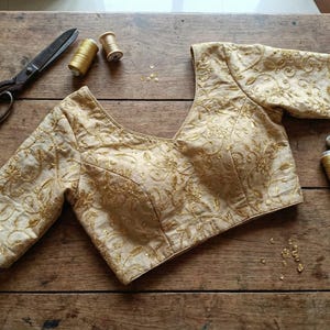 Puede incluir: Una blusa beige bordada con mangas tres cuartos, con intrincados patrones florales dorados. La blusa se muestra sobre una superficie de madera junto con herramientas de costura, incluyendo tijeras, carretes de hilo y una cinta métrica. La estética general sugiere ropa artesanal.