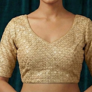 Puede incluir: Una blusa dorada de manga corta con escote en V. La tela tiene un patrón texturizado en forma de escamas y está adornada con pequeñas lentejuelas brillantes. La prenda está diseñada para ser usada como top.