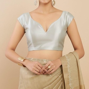 May include: A light silver-colored blouse with a deep V-neckline and cap sleeves. The blouse is paired with a gold-colored saree. The model is wearing gold earrings, a gold bracelet, and a ring.