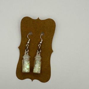 May include: A pair of dangle earrings featuring miniature clear glass bottles filled with iridescent white glitter. The bottles are attached to silver-toned metal fishhook ear wires and are displayed on a brown scalloped cardboard backing.