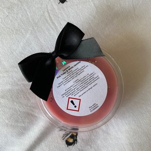 May include: A pink wax melt in a clear container with a black bow on top. The label reads "Leonora Pink Tulips & White Jasmine" and includes a warning about the product being harmful to aquatic life. The label also lists the ingredients and contact information for the manufacturer.