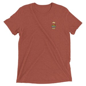 May include: A rust-colored t-shirt featuring a small embroidered burger design on the chest. The burger includes a bun, lettuce, tomato, patty, cheese, and onion. The shirt has short sleeves and a crew neck.