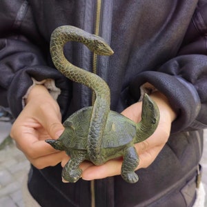 Op de afbeelding: Een bronzen sculptuur van een schildpad met een slang. De schildpad heeft een gedetailleerd schildpatroon en de slang is om het lichaam van de schildpad gewikkeld. De sculptuur heeft een verweerde, antieke uitstraling.