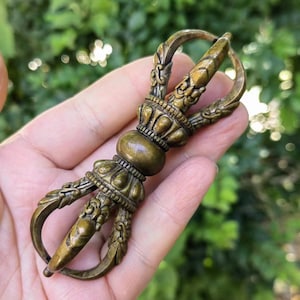 May include: A bronze-colored Vajra, a ritual object, is held in a hand. The Vajra features intricate details, including curved prongs and ornate floral designs. The object has a cylindrical center and is likely made of metal.