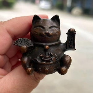May include: Bronze Maneki Neko figurine, a traditional Japanese lucky cat. The cat is depicted with one paw raised, holding a fan and a scroll. The figurine has a smiling expression and is holding a bowl of coins. The cat is approximately 3 inches tall.