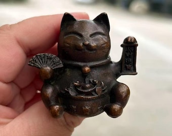 May include: Bronze Maneki Neko figurine, a traditional Japanese lucky cat. The cat is depicted with one paw raised, holding a fan and a scroll. The figurine has a smiling expression and is holding a bowl of coins. The cat is approximately 3 inches tall.