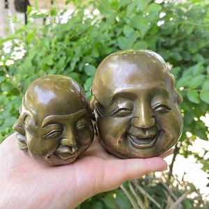 Op de afbeelding: Twee bronzen Boeddha-hoofdsculpturen. De ene sculptuur heeft drie gezichten en de andere heeft één lachend gezicht. De sculpturen worden in een hand gehouden tegen een achtergrond van groen gebladerte.