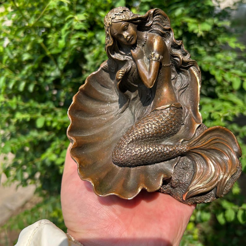 May include: A bronze-toned decorative figurine featuring a mermaid with long flowing hair resting inside a large, open seashell. The mermaid has a detailed scaly tail and is positioned with one hand near her face, set against a textured shell base.